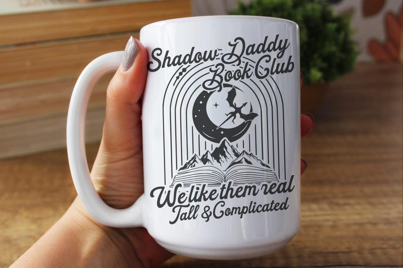 May include: White ceramic mug with a black graphic design featuring a crescent moon, a dragon, and a book. The text reads "Shadow Daddy Book Club We like them real Tall & Complicated".