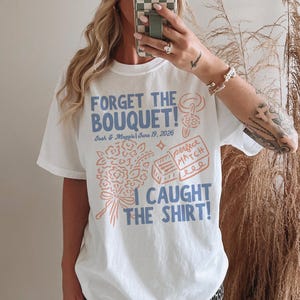 May include: White t-shirt with a blue and peach graphic design. The text reads "FORGET THE BOUQUET!" with the names "Josh & Maggie" and the date "June 19, 2026". Below the text is a bouquet and a matchbox design. The phrase "I CAUGHT THE SHIRT!" is at the bottom.