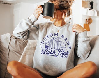 May include: A light gray sweatshirt with a navy blue graphic that says "Custom Name Book Club est. 2025". The graphic includes illustrations of books, flowers, and a teacup. The person is holding a black mug.