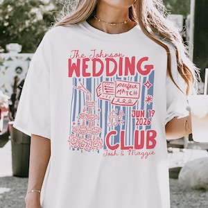 Op de afbeelding: Wit T-shirt met een rood en blauw grafisch ontwerp. Het ontwerp bevat de tekst "The Johnson Wedding Club" en de datum "JUN 19 2026". Illustraties zijn onder meer champagneglazen, een taart en muzieknoten.