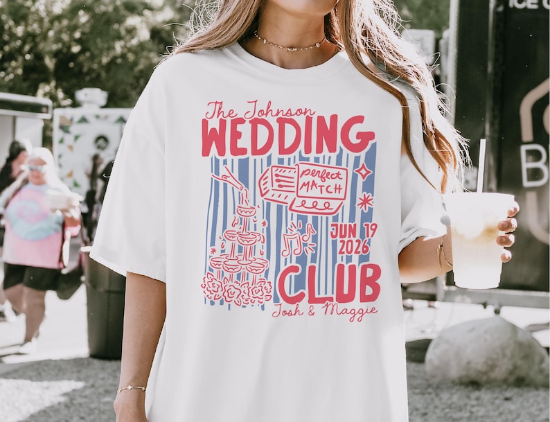 Op de afbeelding: Wit T-shirt met een rood en blauw grafisch ontwerp. Het ontwerp bevat de tekst "The Johnson Wedding Club" en de datum "JUN 19 2026". Illustraties zijn onder meer champagneglazen, een taart en muzieknoten.