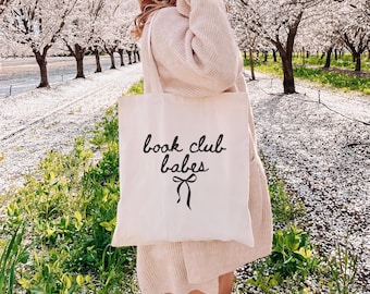 May include: Cream-colored tote bag with the text "book club babes" in black script, accented by a bow. The bag is carried outdoors, with a background of flowering trees and green grass.