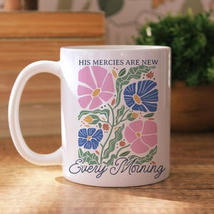 K&ouml;nnte beinhalten: Wei&szlig;e Keramiktasse mit floralem Design mit rosa und blauen Blumen, gr&uuml;nen Bl&auml;ttern und dem Text "HIS MERCIES ARE NEW Every Morning". Die Tasse hat einen gebogenen Henkel und steht auf einer Holzoberfl&auml;che.