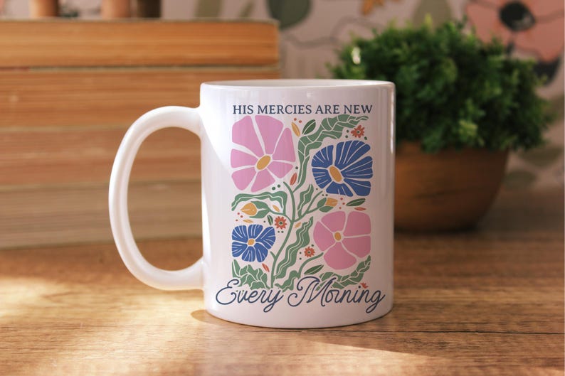 K&ouml;nnte beinhalten: Wei&szlig;e Keramiktasse mit floralem Design mit rosa und blauen Blumen, gr&uuml;nen Bl&auml;ttern und dem Text "HIS MERCIES ARE NEW Every Morning". Die Tasse hat einen gebogenen Henkel und steht auf einer Holzoberfl&auml;che.