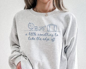 Könnte beinhalten: Hellgraues Sweatshirt mit blauem Reise-Themen-Motiv. Das Design umfasst einen Globus, einen Reisepass, ein Flugzeug und einen Koffer mit dem Schriftzug "a little something to take the edge off".