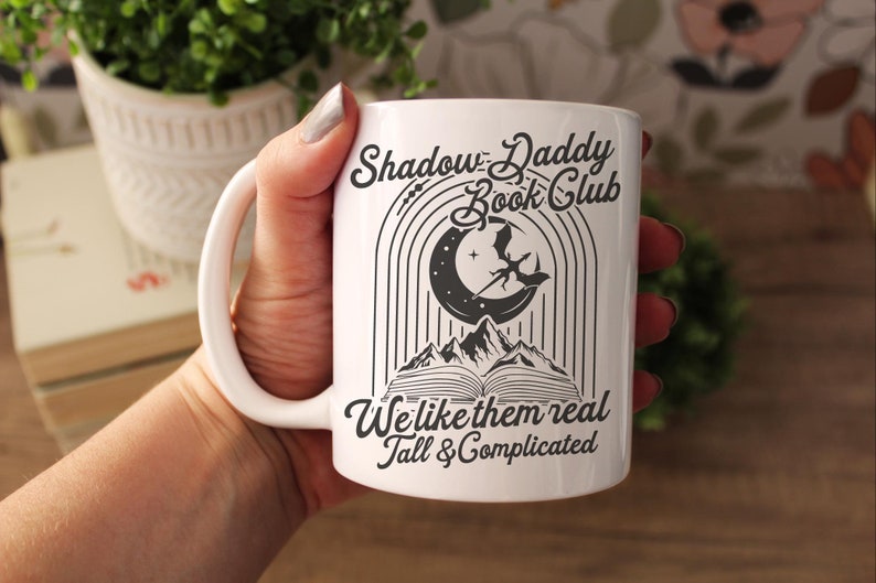 May include: White ceramic mug with a black design that says "Shadow Daddy Book Club" with a crescent moon, a dragon, and mountains. The text below the design says "We like them real tall & complicated."