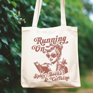 May include: A white tote bag with a red graphic of a woman reading a book and holding a drink. The text on the bag reads "Running on Spicy Books & Caffeine".