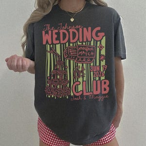 Op de afbeelding: Donkergrijs t-shirt met een rood en geel grafisch ontwerp. Het ontwerp bevat de woorden "The Johnson WEDDING CLUB" en "Jun 19 2026". Het shirt heeft ook illustraties van een taart, champagne toren en muzieknoten.