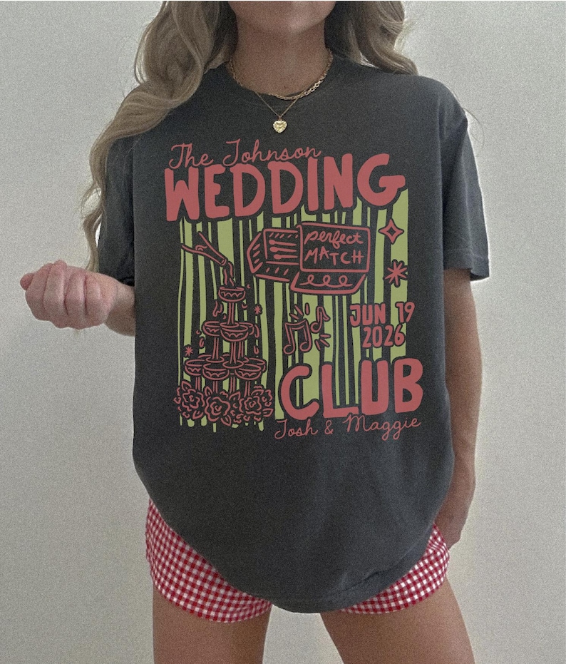 Op de afbeelding: Donkergrijs t-shirt met een rood en geel grafisch ontwerp. Het ontwerp bevat de woorden "The Johnson WEDDING CLUB" en "Jun 19 2026". Het shirt heeft ook illustraties van een taart, champagne toren en muzieknoten.