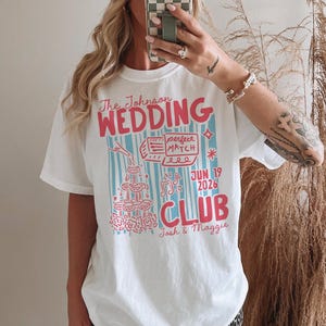 Op de afbeelding: Wit T-shirt met een kleurrijk grafisch ontwerp. Het ontwerp bevat de tekst "The Johnson WEDDING CLUB" in roze en blauw, samen met de datum "JUN 19 2026" en de namen "Josh & Maggie". Het shirt heeft een casual, oversized pasvorm.