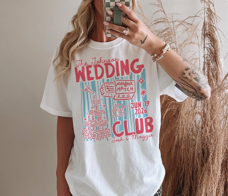 Op de afbeelding: Wit T-shirt met een kleurrijk grafisch ontwerp. Het ontwerp bevat de tekst "The Johnson WEDDING CLUB" in roze en blauw, samen met de datum "JUN 19 2026" en de namen "Josh & Maggie". Het shirt heeft een casual, oversized pasvorm.