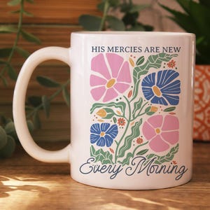 K&ouml;nnte beinhalten: Wei&szlig;e Keramiktasse mit floralem Design. Das Design umfasst rosa und blaue Blumen, gr&uuml;ne Bl&auml;tter und den Text "HIS MERCIES ARE NEW Every Morning". Die Tasse hat einen gebogenen Henkel und steht auf einer Holzoberfl&auml;che.