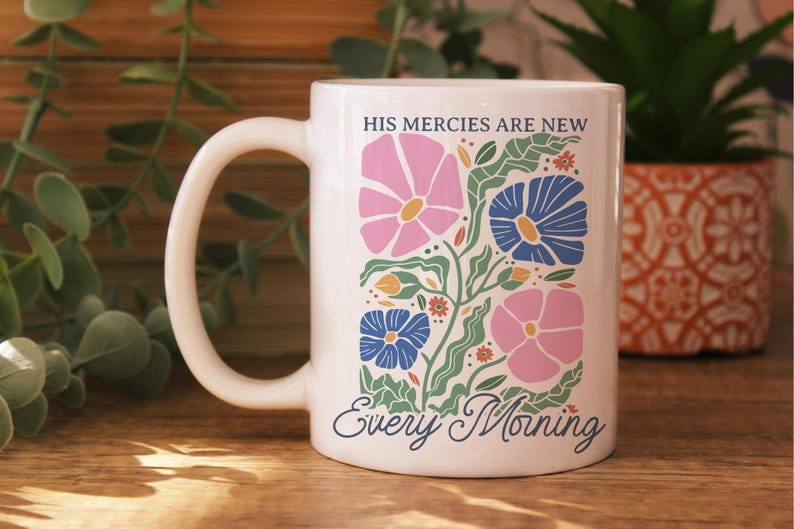 K&ouml;nnte beinhalten: Wei&szlig;e Keramiktasse mit floralem Design. Das Design umfasst rosa und blaue Blumen, gr&uuml;ne Bl&auml;tter und den Text "HIS MERCIES ARE NEW Every Morning". Die Tasse hat einen gebogenen Henkel und steht auf einer Holzoberfl&auml;che.