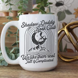 May include: White ceramic mug with a black and white design. The mug features a crescent moon, a dragon, and an open book. The text on the mug reads "Shadow Daddy Book Club We like them real Tall & Complicated".