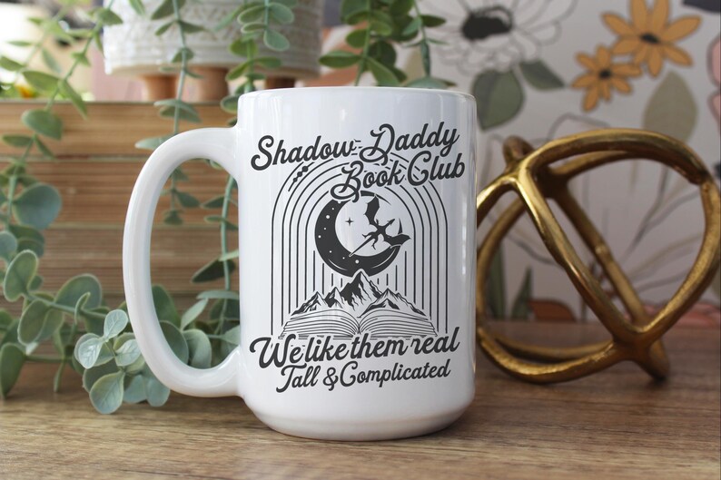 May include: White ceramic mug with a black and white design. The mug features a crescent moon, a dragon, and an open book. The text on the mug reads "Shadow Daddy Book Club We like them real Tall & Complicated".