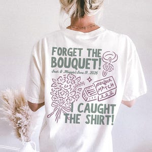 May include: Cream-colored t-shirt with a graphic design in green and purple. The text reads "FORGET THE BOUQUET!" with the names "Josh & Maggie" and the date "June 19, 2026". Below the text is a bouquet illustration and the words "I CAUGHT THE SHIRT!"