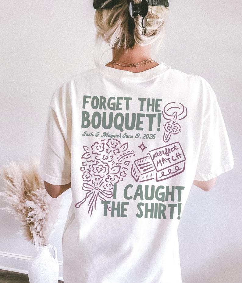 May include: Cream-colored t-shirt with a graphic design in green and purple. The text reads "FORGET THE BOUQUET!" with the names "Josh & Maggie" and the date "June 19, 2026". Below the text is a bouquet illustration and the words "I CAUGHT THE SHIRT!"