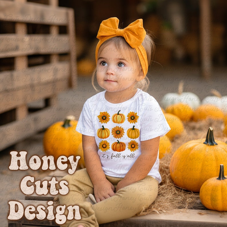 May include: A white t-shirt with a design of orange pumpkins and yellow sunflowers. The text "it's fall y'all" is printed on the shirt. A young child is wearing the shirt and sitting on a bale of hay.