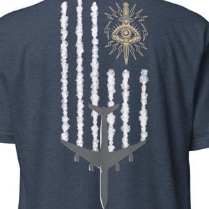 Puede incluir: Camiseta azul marino con un diseño estilizado. El diseño incluye líneas verticales blancas que se asemejan a estelas de condensación, un avión plateado y un emblema dorado con un ojo, una daga y rayos. La camiseta está hecha de un material suave.