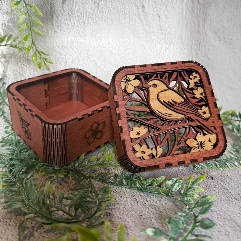 May include: A wooden box with a hinged lid, featuring a laser-cut design of a bird perched on a branch with flowers. The box is open, revealing the interior. The wood has a warm, reddish-brown tone, and the design is intricate.