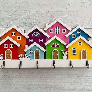 May include: A colorful wooden key holder shaped like a row of houses. Each house is painted a different color, including red, orange, purple, blue, green, and yellow. The houses have white trim and windows. There are five hooks on the bottom of the key holder.