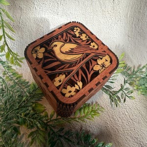 May include: A square wooden box with a detailed laser-cut design. The top features a bird perched on a branch with flowers, all in a light brown color against a darker brown background. The box is surrounded by green foliage.