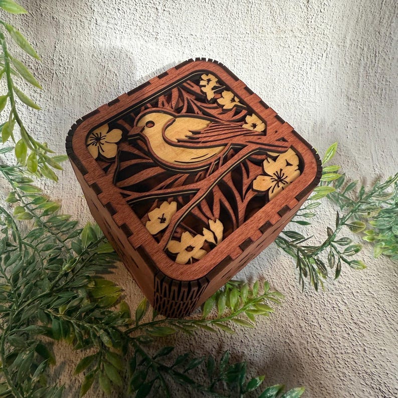May include: A square wooden box with a detailed laser-cut design. The top features a bird perched on a branch with flowers, all in a light brown color against a darker brown background. The box is surrounded by green foliage.