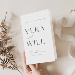 Könnte beinhalten: Eine minimalistische Hochzeitseinladung mit den Namen "VERA und WILL". Die Einladung wird von einer Hand gehalten, mit einem getrockneten Farn und weißem Band im Hintergrund. Das Datum ist der 14. April 2024.