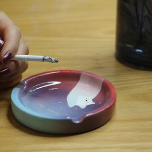 Puede incluir: Un cenicero de cerámica con un degradado de colores, incluyendo rojo, morado y azul. El cenicero tiene un diseño único con una forma blanca y una pequeña muesca para sostener un cigarrillo. Un cigarrillo encendido está encima.
