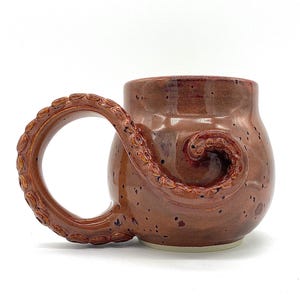 May include: A brown ceramic mug with a unique handle shaped like an octopus tentacle. The mug has a speckled finish and a reddish-brown glaze. The handle curves into the body of the mug, adding a distinctive design element. The mug is suitable for hot or cold beverages.