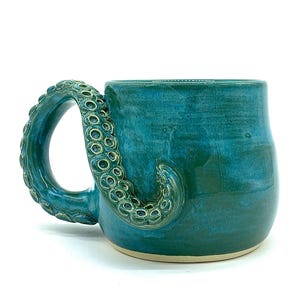 May include: A ceramic mug with a glossy teal glaze and a handle sculpted in the shape of an octopus tentacle. The tentacle wraps around the side of the mug, featuring detailed circular suction cups along its length. The base is unglazed and beige.