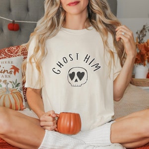Puede incluir: Camiseta color crema con el texto "GHOST HIM" sobre una calavera con ojos en forma de corazón. La persona sostiene una taza naranja con forma de calabaza. La camiseta forma parte de una colección temática de Halloween o otoño.