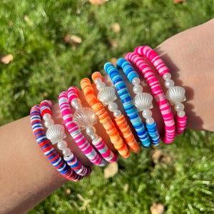 May include: A stack of colorful bracelets with white beads and shell charms. The bracelets are made of layered, striped, and solid colored beads.