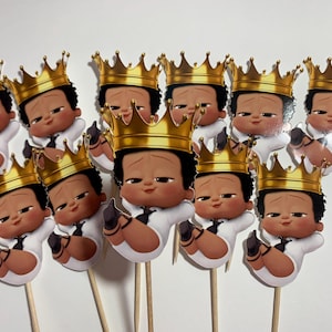 May include: A collection of baby-themed party decorations featuring cartoon images of a baby wearing a gold crown, white suit, and black tie. The baby images are attached to wooden sticks.