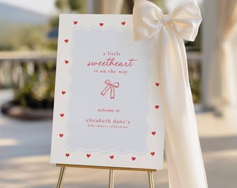 May include: A baby shower welcome sign on a gold easel. The sign is white with a scalloped border and red hearts. Text reads "a little sweetheart is on the way" and "welcome to elizabeth dane's baby shower celebration". A large cream bow adorns the side.