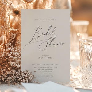 May include: A cream-colored invitation card with the words "Bridal Shower" in elegant script. The card includes details such as the honoree's name, date, time, and location. The card is set on a table with a glass and dried flowers.