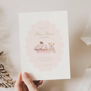 May include: A cream-colored invitation card with a pink scalloped border. The card features the text "Blair Bear's First Birthday" and an illustration of a teddy bear and cakes. The text "Saturday, 02 July 2010" is also included.