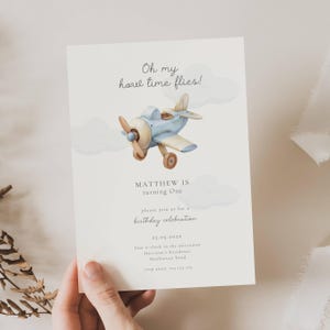 May include: A light cream-colored birthday invitation featuring a watercolor illustration of a vintage airplane in shades of blue, tan, and brown. The text "Oh my how time flies!" and "MATTHEW IS turning One" are displayed.