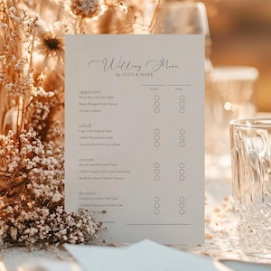 May include: A wedding menu card with the text "Wedding Menu by LUCY & MARK" in elegant script. The menu lists appetizers, salads, entrees, and desserts with space for guest names. The card is a soft cream color.