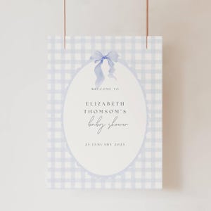 May include: A baby shower sign with a blue gingham pattern background. The sign features an oval shape with the words "Welcome to Elizabeth Thomson's baby shower" and the date "25 January 2025". A blue bow is at the top.