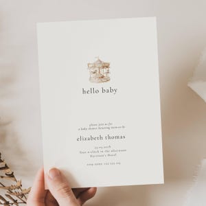 May include: A cream-colored baby shower invitation with a carousel illustration at the top. The text "hello baby" is centered below the carousel. Additional text includes the event details and RSVP information.