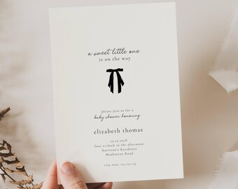Könnte beinhalten: Cremefarbene Babyparty-Einladung mit schwarzem Text und einer schwarzen Schleifenillustration. Der Text lautet "a sweet little one is on the way" und lädt Gäste ein, Elizabeth Thomas zu ehren. Enthält Datum, Uhrzeit und Ort.