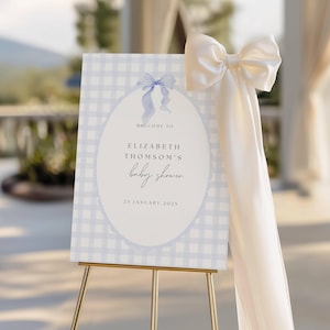 May include: A baby shower welcome sign on a gold easel. The sign has a light blue gingham pattern background with an oval in the centre. The text reads "Welcome to Elizabeth Thomson's baby shower, 25 January 2025". A cream-coloured bow is attached.