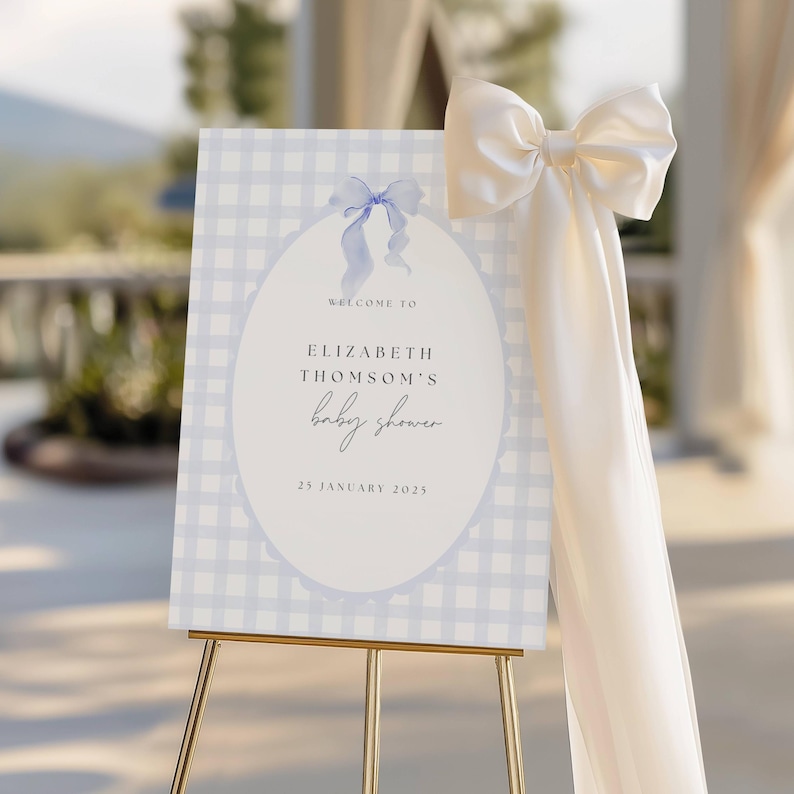 May include: A baby shower welcome sign on a gold easel. The sign has a light blue gingham pattern background with an oval in the center. The text reads "Welcome to Elizabeth Thomson's baby shower, 25 January 2025". A cream-colored bow is attached.