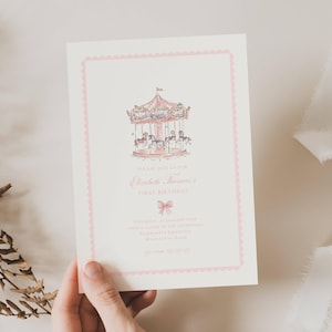 Op de afbeelding: Een lichtroze uitnodigingskaart met een carrousel illustratie. De kaart bevat de tekst "Please join us for Elizabeth Thomas's First Birthday" en evenementdetails. De kaart is omlijst met een geschulpte roze rand.