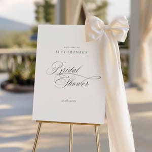 May include: A white sign on a gold easel, welcoming guests to Lucy Thomas's Bridal Shower. The sign features elegant script and is adorned with a large, cream-colored bow and flowing ribbon. The date 25.05.202X is printed at the bottom.