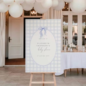 May include: A baby shower welcome sign on a wooden easel. The sign has a light blue gingham pattern background with an oval shape in the center. The oval contains the text "Welcome to Elizabeth Thomson's baby shower" and the date.