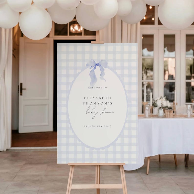 May include: A baby shower welcome sign on a wooden easel. The sign has a light blue gingham pattern background with an oval shape in the center. The oval contains the text "Welcome to Elizabeth Thomson's baby shower" and the date.
