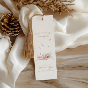 May include: A cream-colored bookmark with a gold tassel. The bookmark features the text "Elizabeth's new chapter" and "Thank You" with floral and butterfly illustrations. The bookmark is hanging on a white cloth.