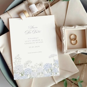 May include: A white Save the Date card featuring blue hydrangea illustrations, the names Lucy Ingrid and Charlie Evans, and the date 25.05.202X. The card is displayed with cream envelopes, two small spools of ribbon, and a small ornate frame with two gold rings.